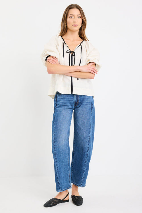Woman wearing a cream long sleeve linen top with black trim and tie-front detail, designed in an oversized fit with puff sleeves, paired with blue jeans.