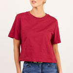 A woman modeling a short-sleeve crewneck t-shirt in a deep red color, paired with blue jeans, against a plain off-white studio background.