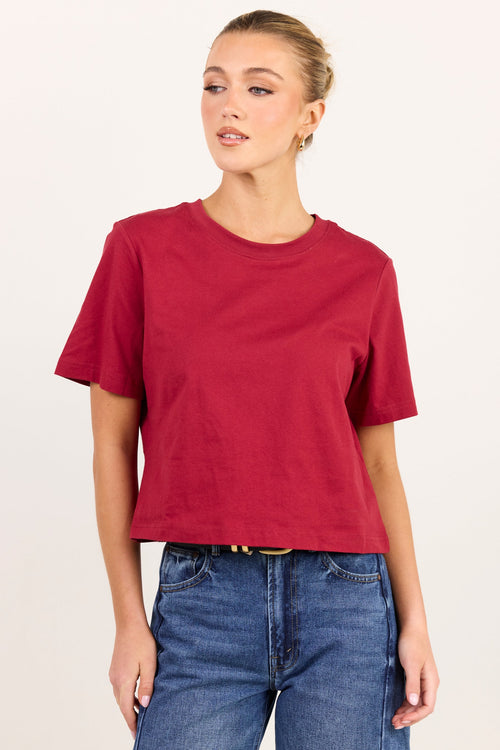 A woman modeling a short-sleeve crewneck t-shirt in a deep red color, paired with blue jeans, against a plain off-white studio background.
