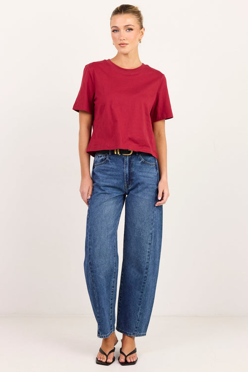 A woman modeling a short-sleeve crewneck t-shirt in deep red and blue jeans, standing against a plain white studio background.