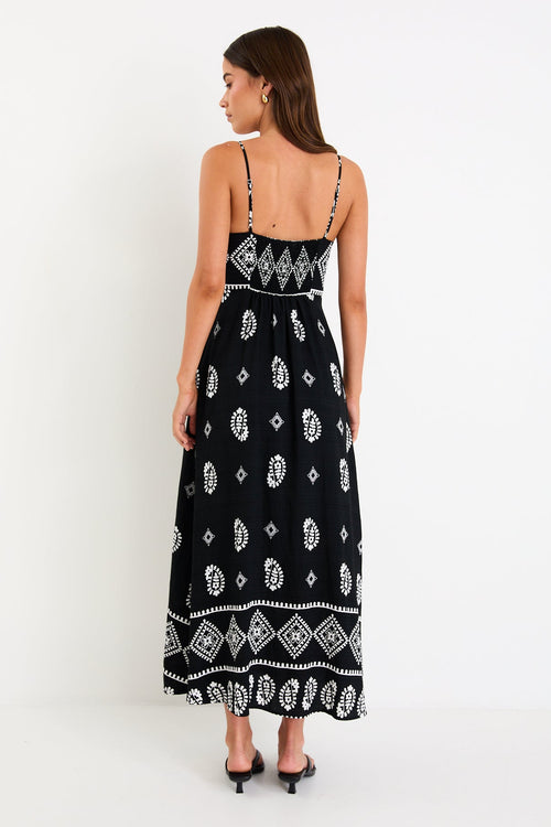 Woman wearing a black maxi dress with fine straps, white paisley and border print, and shirred back detail.