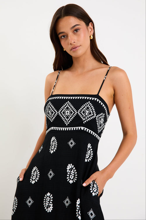 Woman wearing a black maxi dress with fine straps, white paisley and border print, and shirred back detail.