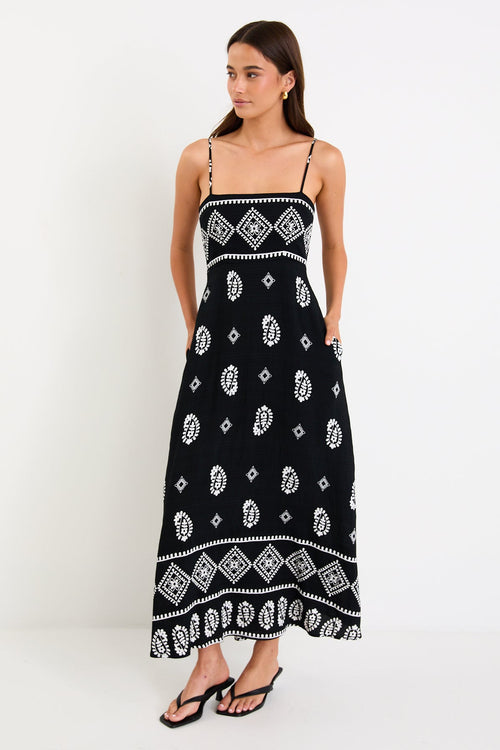 Woman wearing a black maxi dress with fine straps, white paisley and border print, and shirred back detail.