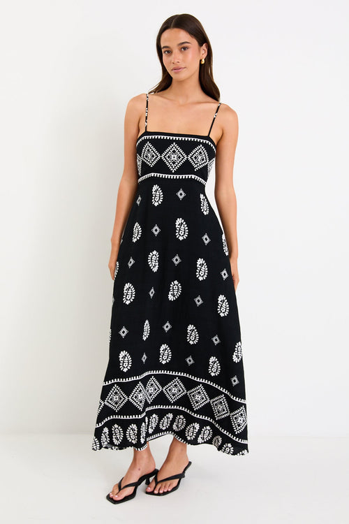 Woman wearing a black maxi dress with fine straps, white paisley and border print, and shirred back detail.