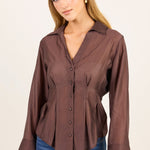A woman modeling a brown long-sleeve button-up blouse with a peplum waist and flared cuffs, paired with blue jeans, against a plain light beige studio background.