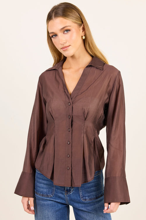 A woman modeling a brown long-sleeve button-up blouse with a peplum waist and flared cuffs, paired with blue jeans, against a plain light beige studio background.