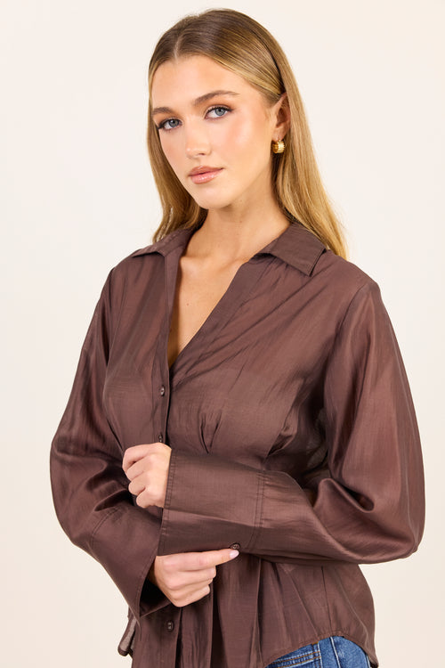 A woman modeling a brown long-sleeve button-up shirt with a gathered waist and wide sleeves, paired with blue jeans against a plain light background.