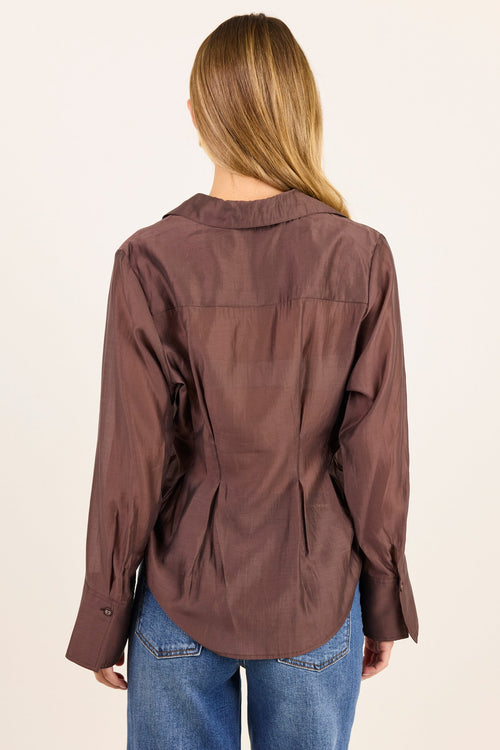 A woman viewed from the back wearing a brown long-sleeve button-up shirt and blue jeans against a plain off-white background.