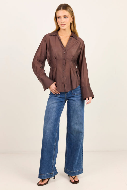 A woman modeling a brown long-sleeve button-up shirt with a shawl collar and flared cuffs, paired with blue wide-leg jeans and brown heeled sandals, against a plain off-white studio background.