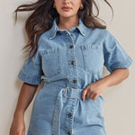 Woman wears a short-sleeve light wash denim mini dress with front buttons, patch pockets, and a matching fabric belt cinched at the waist for a structured, feminine silhouette.