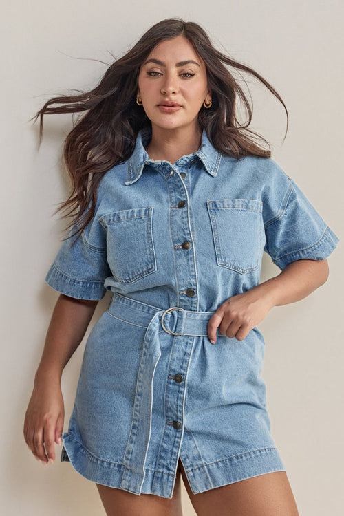 Woman wears a short-sleeve light wash denim mini dress with front buttons, patch pockets, and a matching fabric belt cinched at the waist for a structured, feminine silhouette.