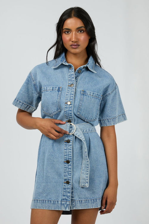 Woman wears a short-sleeve light wash denim mini dress with front buttons, patch pockets, and a matching fabric belt cinched at the waist for a structured, feminine silhouette.