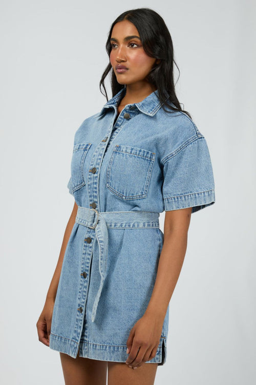 Woman wears a short-sleeve light wash denim mini dress with front buttons, patch pockets, and a matching fabric belt cinched at the waist for a structured, feminine silhouette.