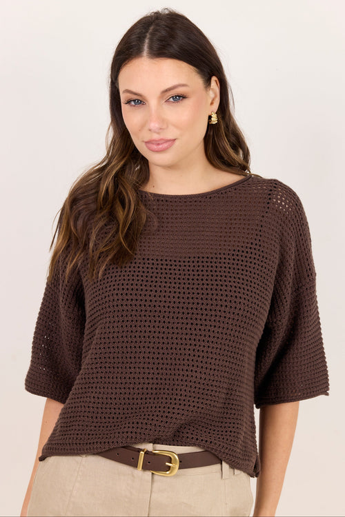 A woman wearing a dark brown open-knit short-sleeve top and beige pants with a brown belt, photographed against a plain light background.