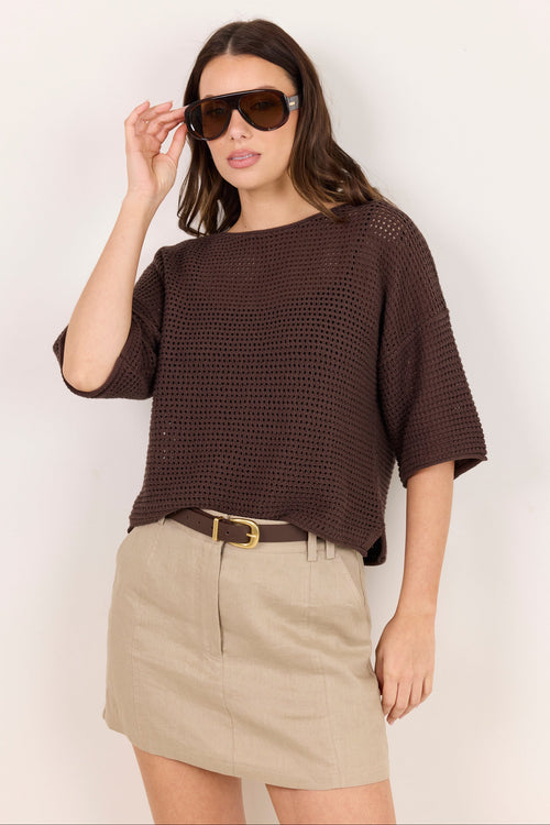 A woman wearing a dark brown open-knit short-sleeve top and a beige mini skirt with a brown belt, standing against a plain off-white background.