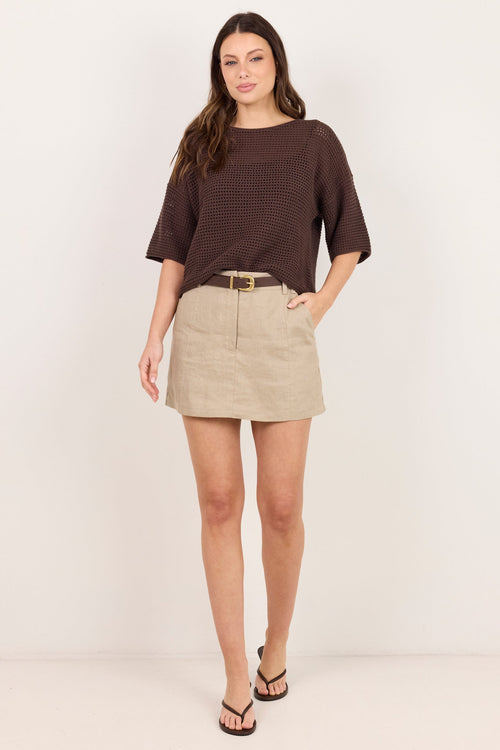 A woman modeling a dark brown short-sleeve knit top and a beige mini skirt with a gold-tone belt, standing against a plain off-white studio background.