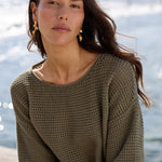 A woman sits on a concrete pier by the water wearing a loose olive green knit top, denim shorts, and black flip-flops.