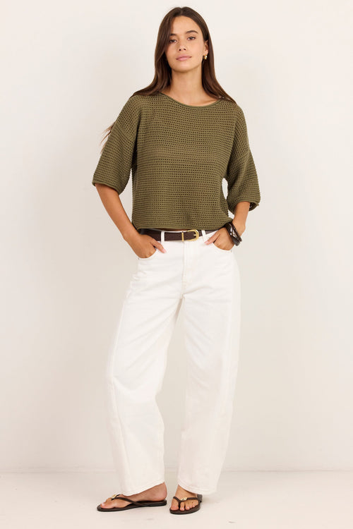A woman modeling a short-sleeve olive green textured knit top with white wide-leg pants and black sandals, standing against a plain off-white studio background.