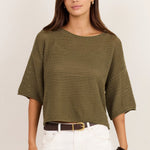 A woman modeling a short-sleeve olive green ribbed knit top with white wide-leg pants, a brown belt, and black sandals, standing against a plain off-white studio background.
