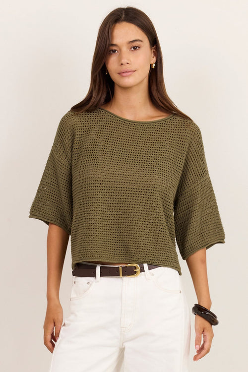 A woman modeling a short-sleeve olive green ribbed knit top with white wide-leg pants, a brown belt, and black sandals, standing against a plain off-white studio background.