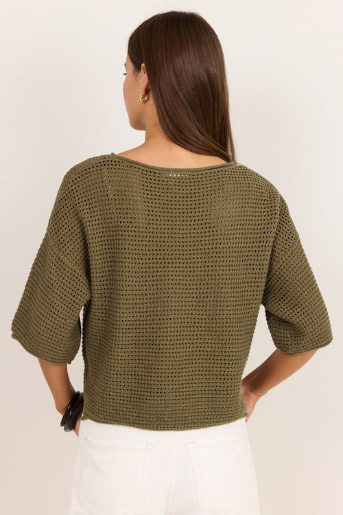 A woman shown from the back wearing a textured olive green knit top with elbow-length sleeves and white pants, against a plain off-white background.