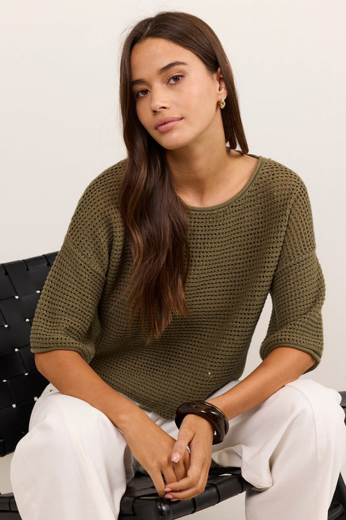 A woman with long dark brown hair sits on a black woven bench, wearing a textured olive green knit top and white pants against a plain off-white background.