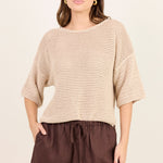 A woman modeling a beige open-knit cropped sweater with elbow-length sleeves and dark brown drawstring shorts against a plain cream background.