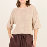 A woman modeling a beige open-knit cropped sweater with elbow-length sleeves and dark brown drawstring shorts against a plain cream background.