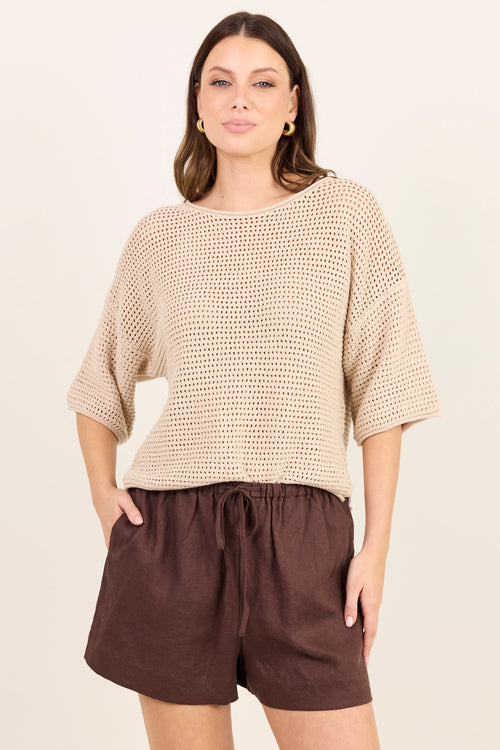 A woman modeling a beige open-knit cropped sweater with elbow-length sleeves and dark brown drawstring shorts against a plain cream background.