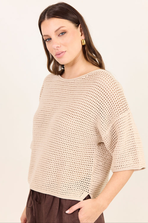 A woman wearing a beige open-knit short-sleeve top and brown pants, photographed against a plain light background.