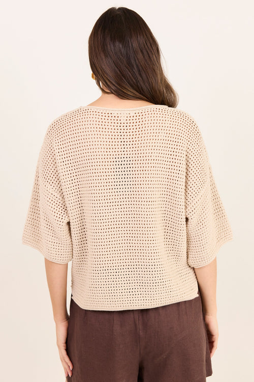 A woman viewed from the back wearing a beige open-knit cropped top with elbow-length sleeves and dark brown pants, against a plain off-white background.
