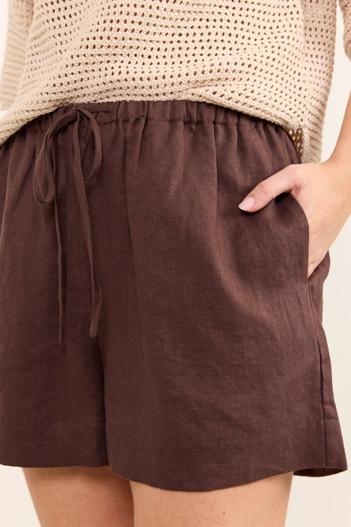 A close-up of a person wearing dark brown linen drawstring shorts with side pockets, paired with a beige knit top, against a plain light background.