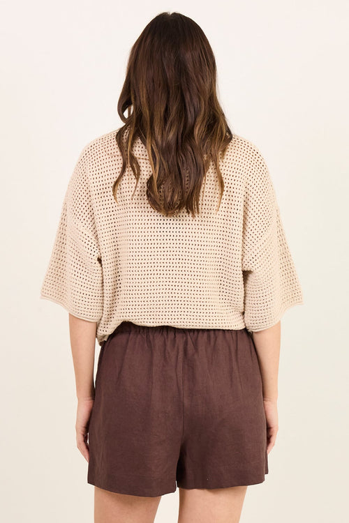 A woman viewed from behind wearing a beige textured short-sleeve top and dark brown shorts, standing against a plain off-white studio background.