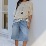 A woman stands outdoors against a white building wall wearing a cream and navy striped knit top, light-wash denim shorts, black flip-flops, and sunglasses.