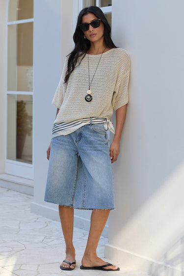 A woman stands outdoors against a white building wall wearing a cream and navy striped knit top, light-wash denim shorts, black flip-flops, and sunglasses.