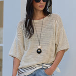 A woman wearing a cream-colored open-knit short-sleeve top, light-wash blue jeans, and black sunglasses, standing against a white stucco wall.