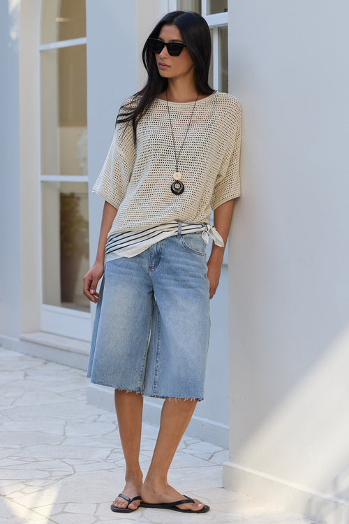 A woman stands outdoors against a white wall wearing a cream knit short-sleeve top, light blue denim culottes, black flip-flops, and sunglasses.