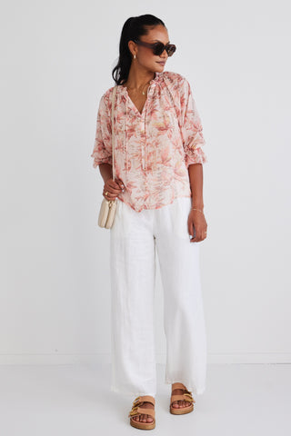 Model wears a floral blouse with white linen pants and brown sunglasses. 