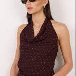 A woman modeling a dark brown halter top with a small white polka dot pattern, paired with blue jeans and black sunglasses, against a plain off-white background.