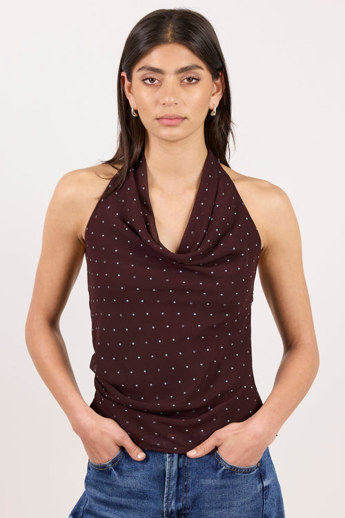 A woman modeling a dark brown halter-neck top with a small white polka dot pattern, paired with blue jeans, against a plain off-white background.