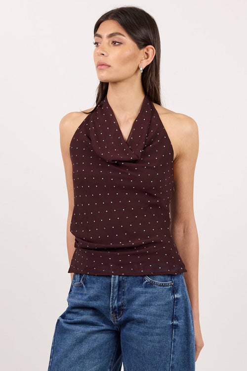 A woman modeling a dark brown halter top with a small white polka dot pattern and blue wide-leg jeans, standing against a plain light gray studio background.