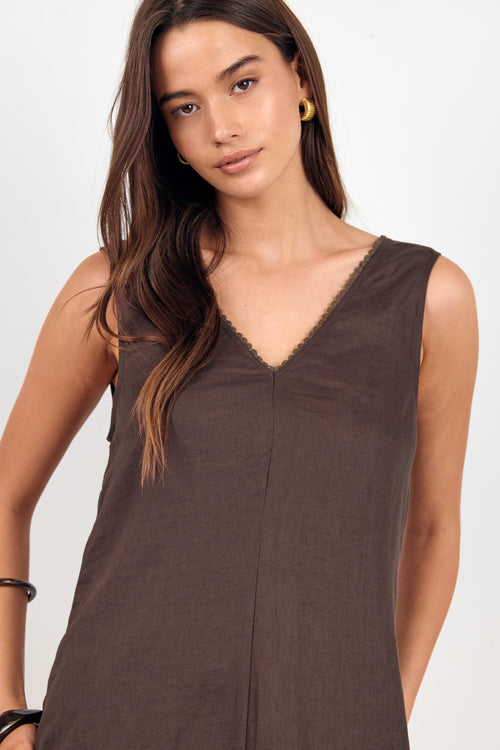 A woman wearing a sleeveless brown V-neck top with delicate scalloped trim along the neckline, photographed against a clean light gray studio background.