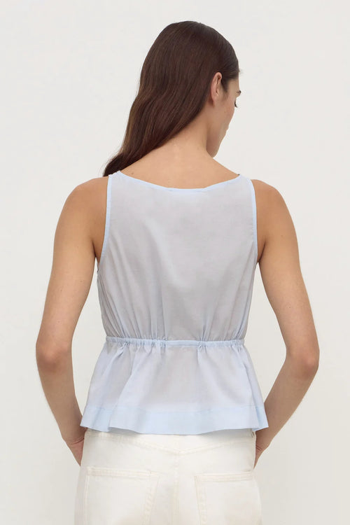 A woman shown from the back wearing a light blue sleeveless peplum top and white pants against a plain white studio background.