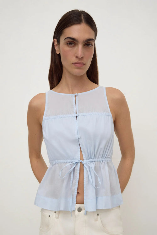 A woman modeling a light blue sleeveless top with a sheer yoke, front button closure, and drawstring waist, paired with off-white pants against a plain light background.
