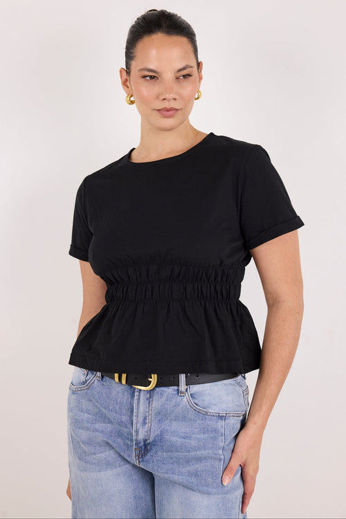 A woman modeling a black short-sleeve top with a gathered smocked waist and peplum hem, paired with light blue jeans and gold hoop earrings, against a plain light gray studio background.