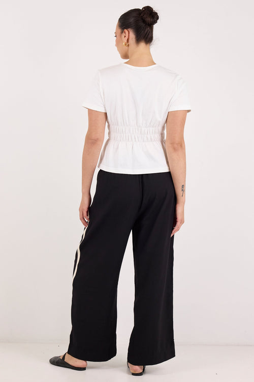 A woman is shown from the back wearing a white short-sleeve cropped top with a smocked waist and black wide-leg pants with white side stripes, set against a plain white studio background.