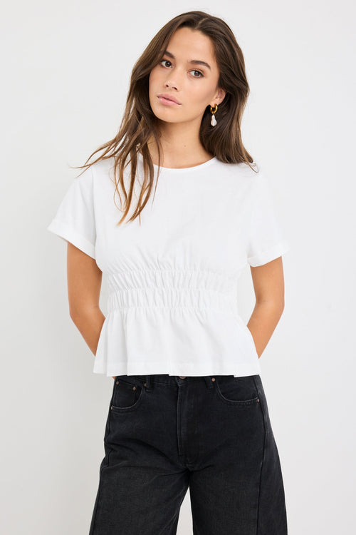 Woman wearing an ivory short sleeve cotton tee with shirred waist detail and a subtle peplum hem, styled with black jeans.