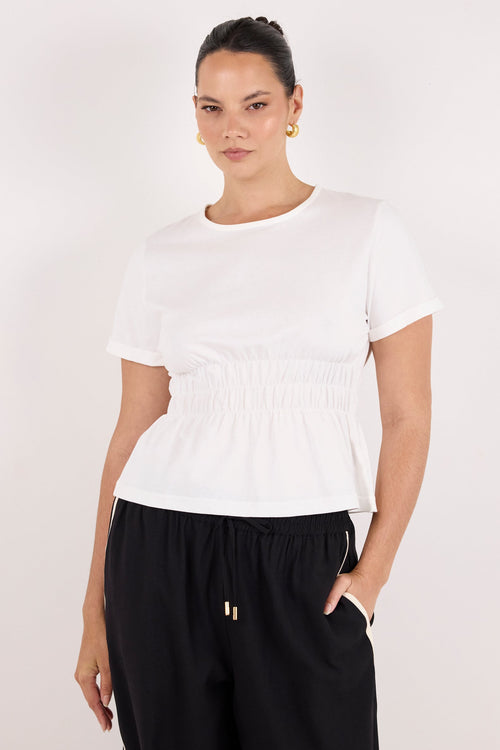 A woman modeling a white short-sleeve crewneck top with a smocked waist, paired with black drawstring pants, against a plain light gray studio background.