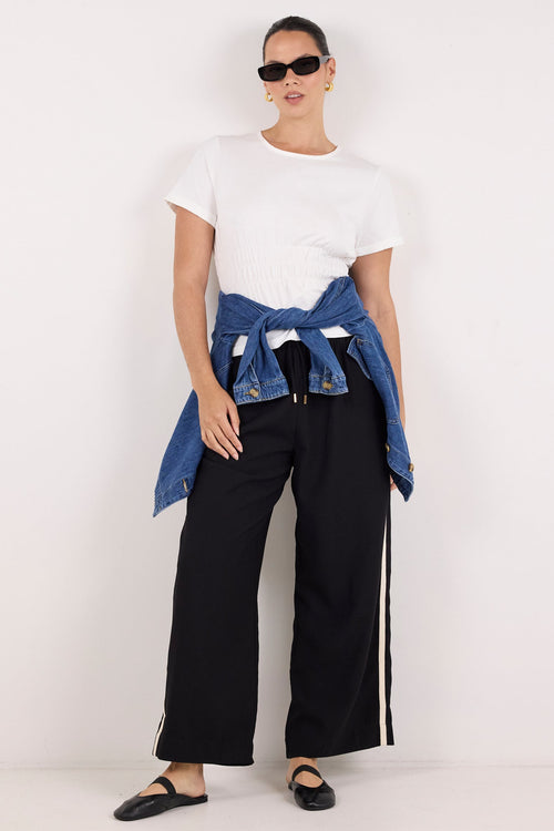 A model stands against a plain white studio background wearing a white short-sleeve t-shirt, black wide-leg pants with white side stripes, black flats, and a blue denim jacket tied around the waist.