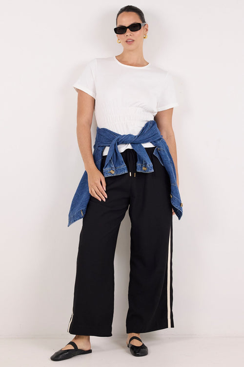 A model stands against a plain white studio background wearing a white short-sleeve t-shirt, black wide-leg pants with white side stripes, black flats, and a blue denim jacket tied around the waist.
